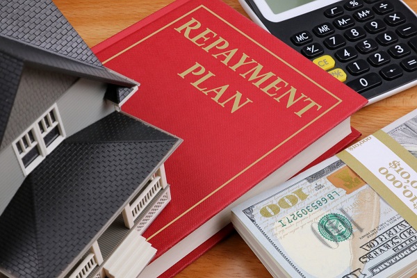 A picture of a book with repayment plan written on the front cover together with a calculator, model house and a stack of 100 dollar bills.